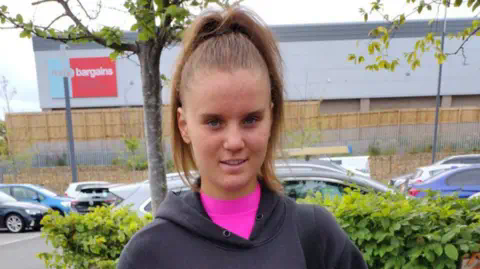 Family handout Holly Newton, photographed in a car park with trees and vehicles behind her, has long dark hair tied up in a pony tail and is wearing a pink shirt under grey hoodie. She is looking directly at the camera